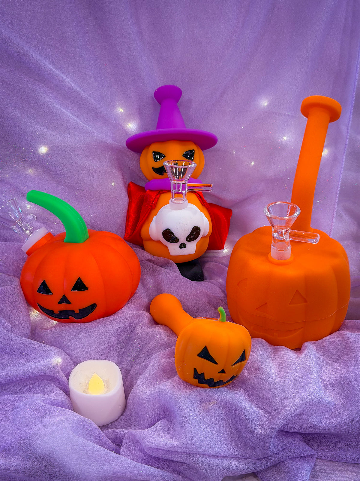 Halloween-themed silicone smoking accessories arranged on draped purple fabric, including orange jack-o'-lantern pipes, a witch figure in a purple hat and red cape holding a skull, and orange and purple smoking accessories.