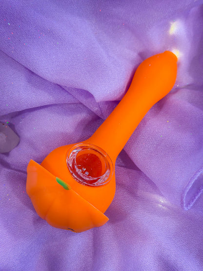 An orange pumpkin-shaped silicone smoking accessory with a clear glass bowl, resting on a purple fabric background.