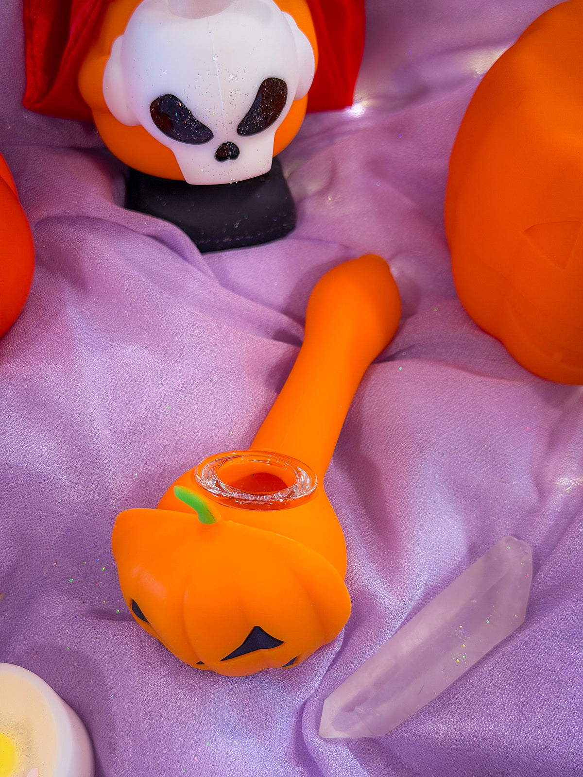 An orange pumpkin-shaped silicone glass pipe with a clear glass bowl, resting on a draped lavender fabric alongside other Halloween-themed props including a jack-o'-lantern figurine and a crystal.