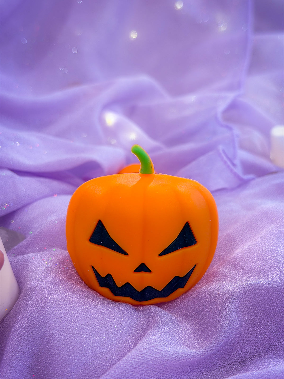 An orange jack-o'-lantern pumpkin pipe with a carved black face and a green stem, placed on draped purple fabric with a soft, sparkly background.